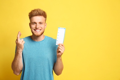 Photo of Portrait of hopeful young man with crossed fingers holding lottery ticket on yellow background, space for text