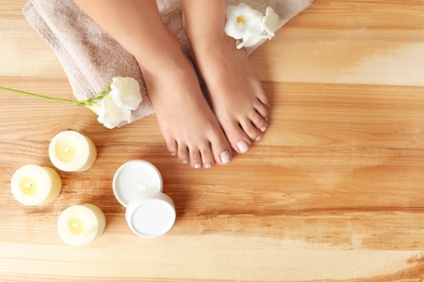 Top view of woman with beautiful feet, candles, flowers and moisturizing cream on wooden floor, space for text. Spa treatment