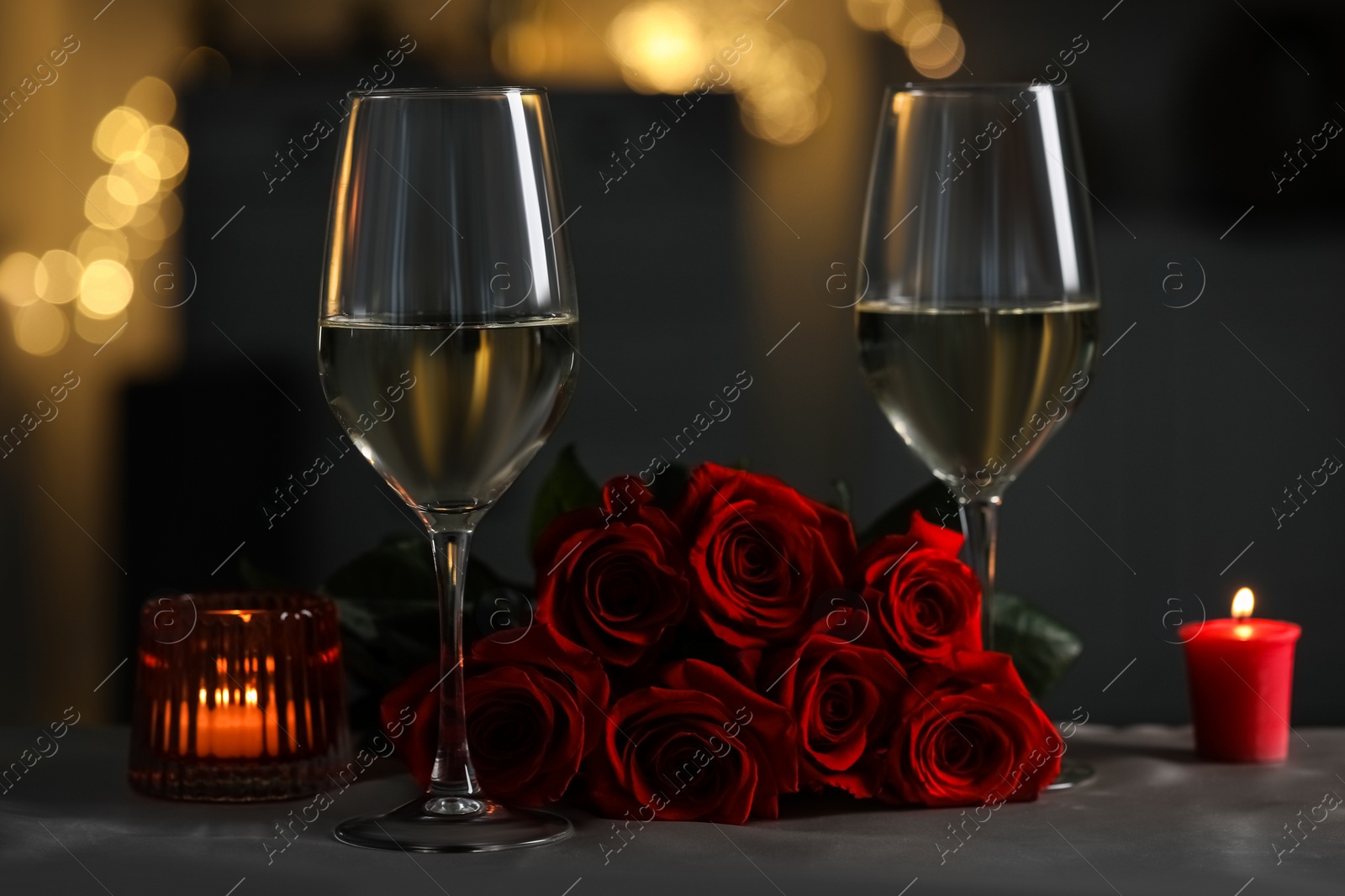 Photo of Romantic table setting with glasses of white wine, burning candles and rose flowers against blurred lights