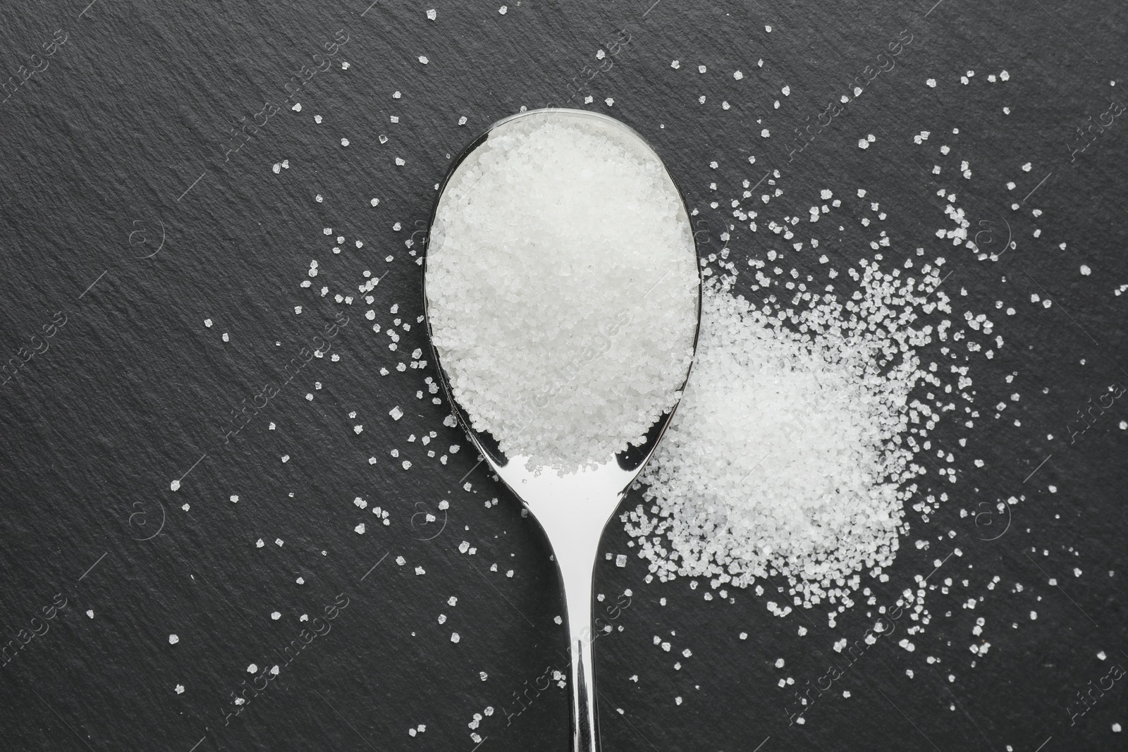 Photo of Granulated sugar and metal spoon on black table, top view