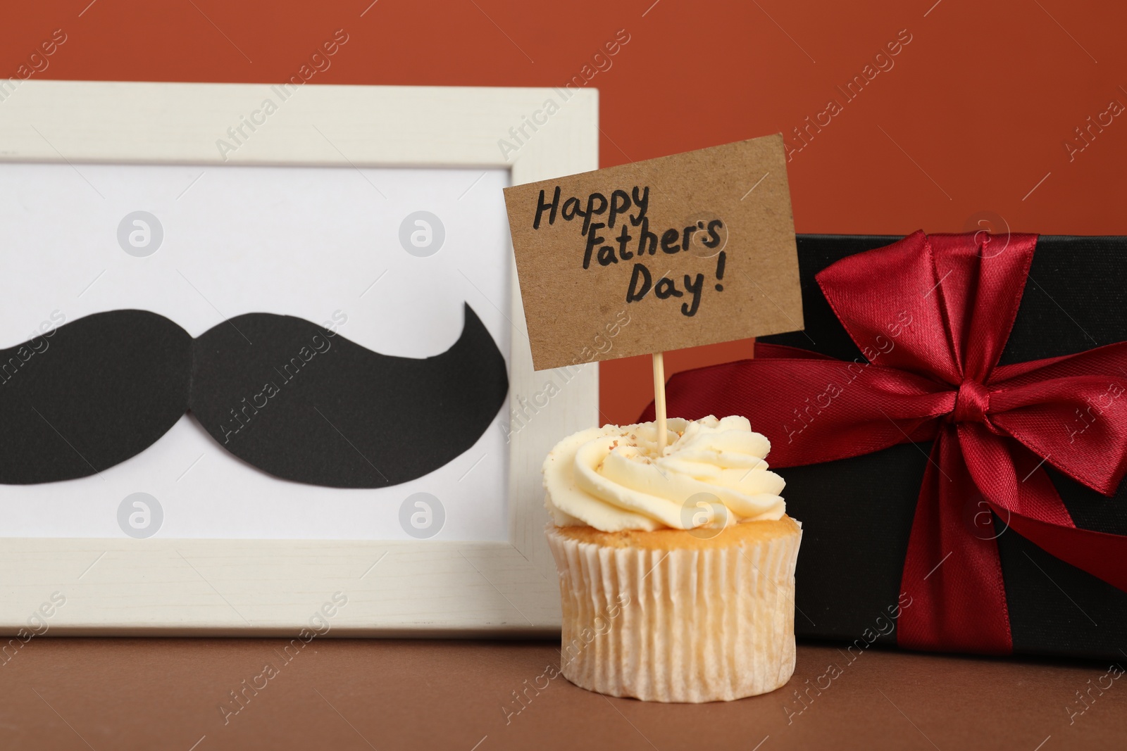 Photo of Happy Father's Day. Tasty cupcake with greetings, gift box and frame with mustache on brown table, closeup