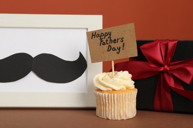 Photo of Happy Father's Day. Tasty cupcake with greetings, gift box and frame with mustache on brown table, closeup