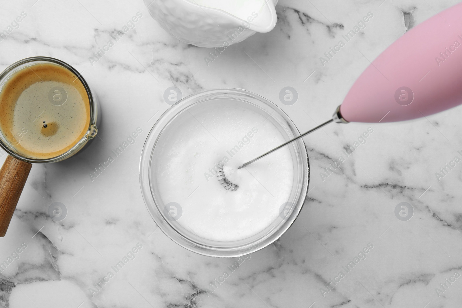 Photo of Whisking milk in glass with mini mixer (milk frother) at white marble table, flat lay