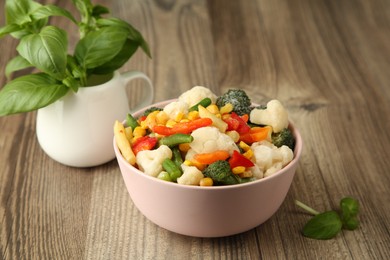 Mix of different frozen vegetables in bowl and basil on wooden table, closeup