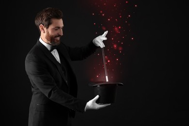 Smiling magician showing trick with wand and top hat on black background