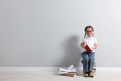 Cute little girl in glasses sitting on stack of books near light grey wall. Space for text