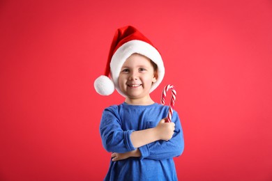 Photo of Cute little boy in Santa Claus hat holding candy cane on red background