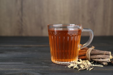 Aromatic licorice tea in cup and dried sticks of licorice root on black wooden table, space for text