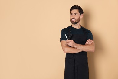 Photo of Smiling hairdresser in apron holding comb and scissors on light brown background, space for text