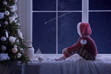 Image of Cute little girl in Santa hat sitting on windowsill and looking at shooting star in beautiful night sky
