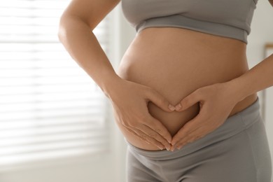 Photo of Pregnant young woman making heart with her hands on belly near window indoors, closeup