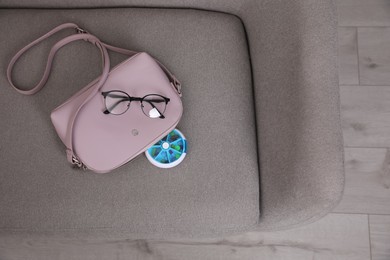 Photo of Pill box with medicaments and glasses near stylish women's bag on sofa indoors, top view. Space for text