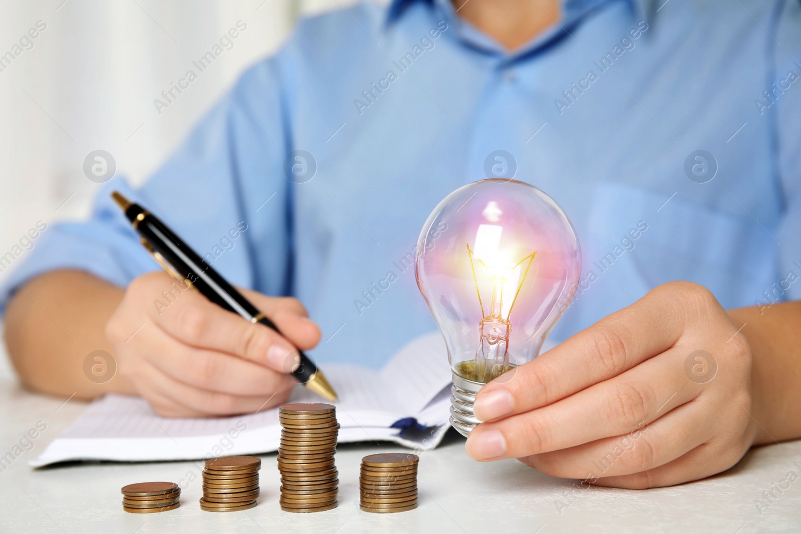 Photo of Woman with light bulb, notebook and coins at white table, closeup. Power saving