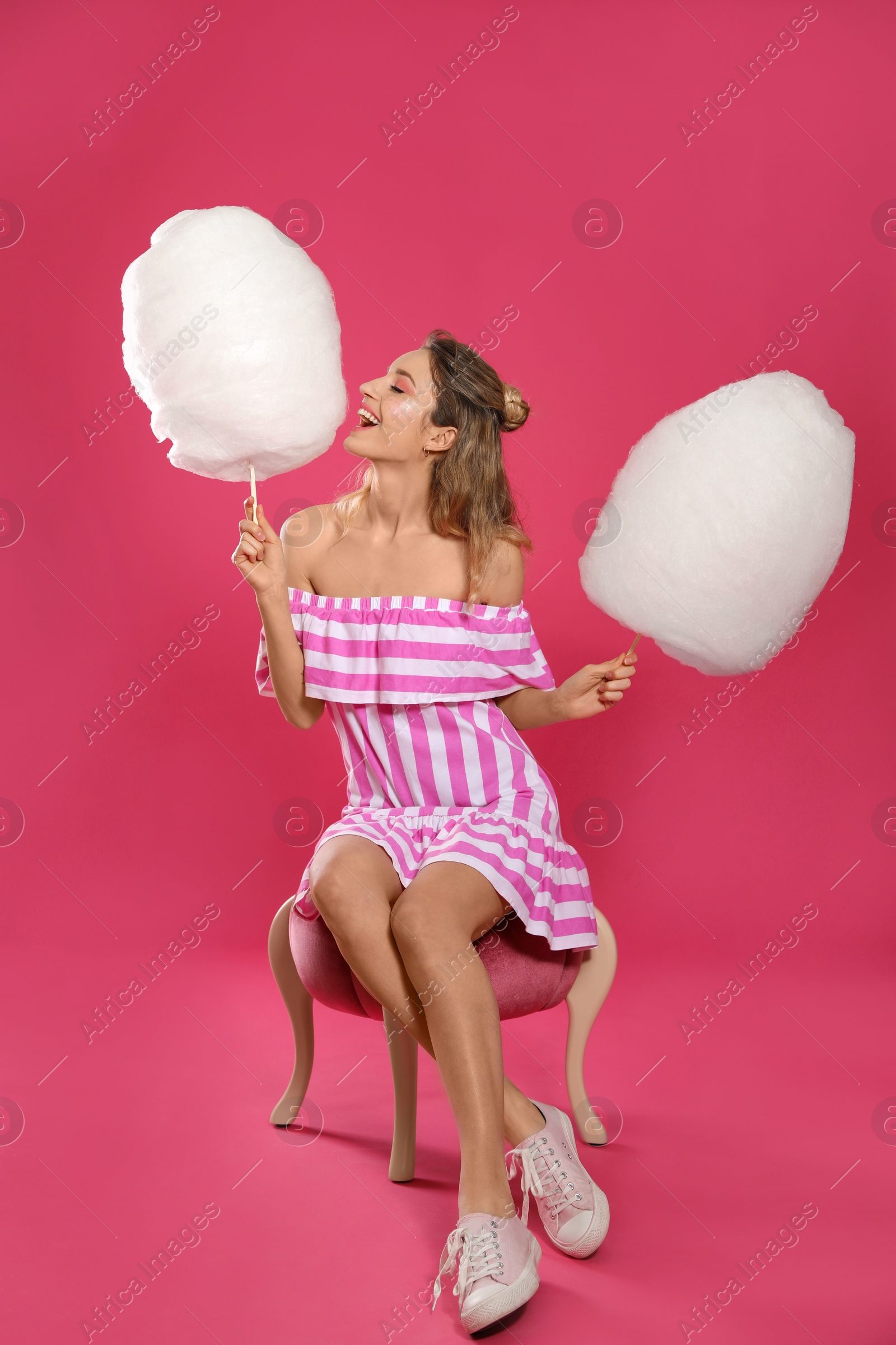 Photo of Full length portrait of young woman with tasty cotton candy sitting on pouf, pink background