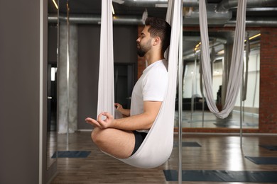 Photo of Young man meditating in fly yoga hammock indoors