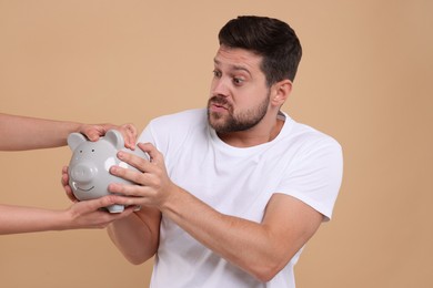 Photo of Woman taking piggy bank from emotional man on beige background. Be careful - fraud