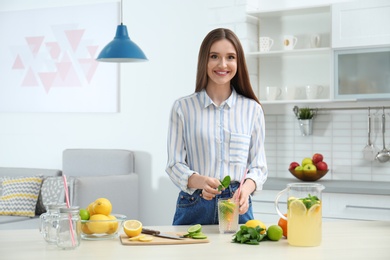 Photo of Beautiful young woman adding mint into glass of natural detox lemonade at table in kitchen