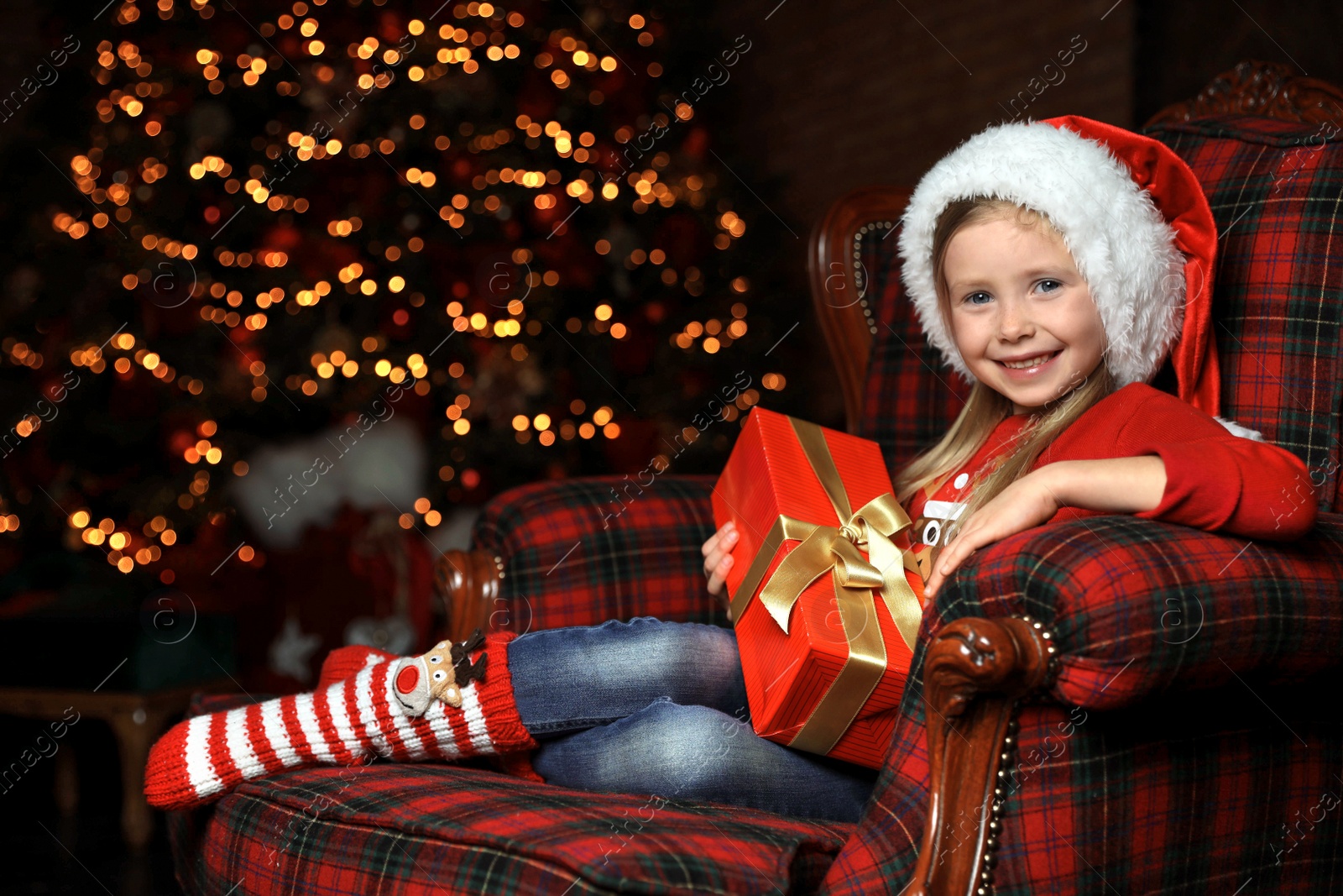 Photo of Cute little child with Christmas gift sitting in armchair at home