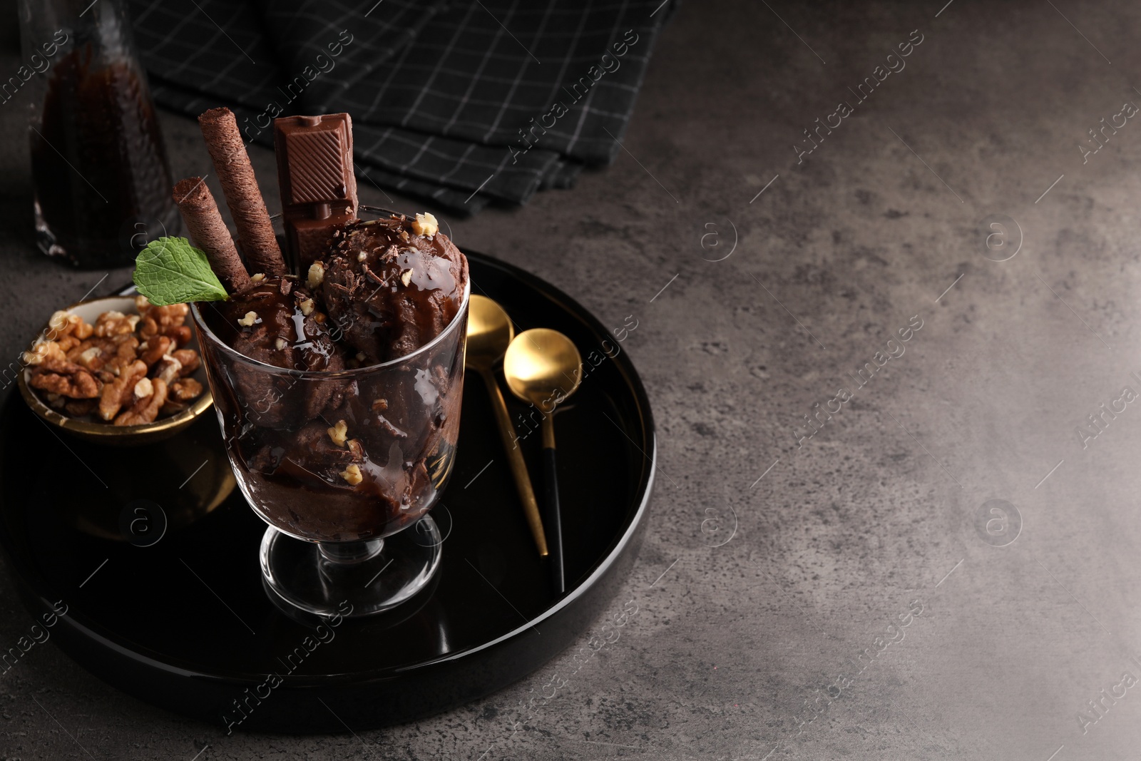 Photo of Tasty chocolate ice cream with sauce, nuts and wafer rolls in glass dessert bowl on dark grey table. Space for text