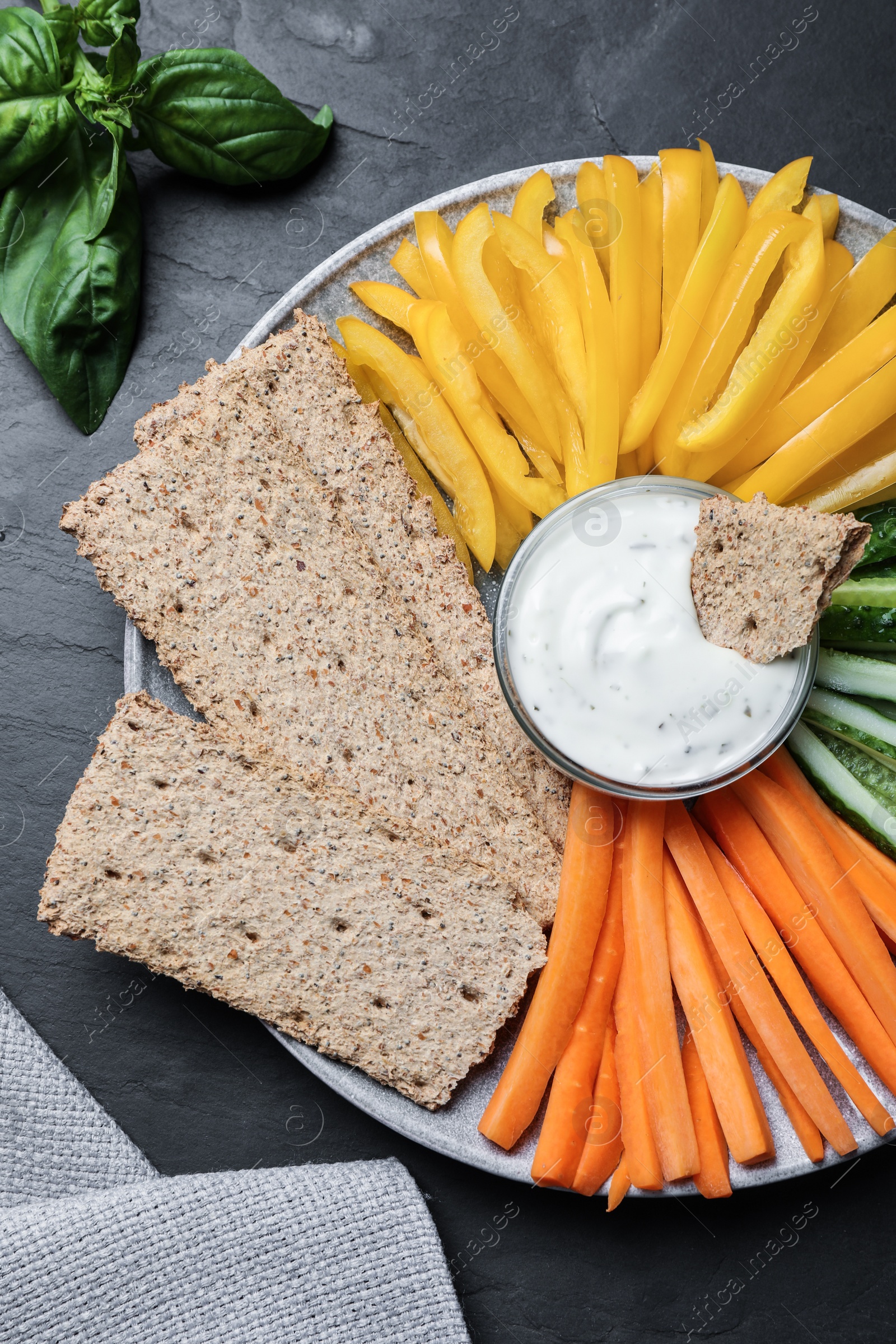 Photo of Plate with crispbreads, vegetable sticks and dip sauce on black table, top view