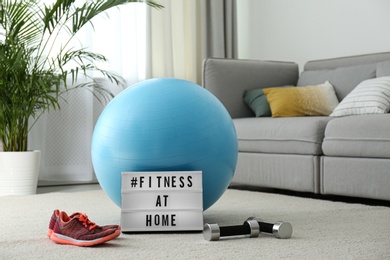 Photo of Sport equipment and lightbox with hashtag FITNESS AT HOME on floor indoors. Message to promote self-isolation during COVID‑19 pandemic