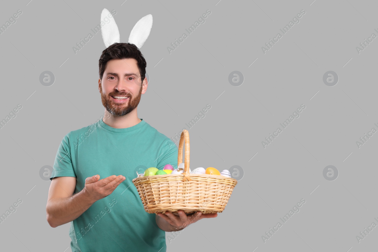 Photo of Portrait of happy man in cute bunny ears headband holding basket with Easter eggs on light grey background. Space for text