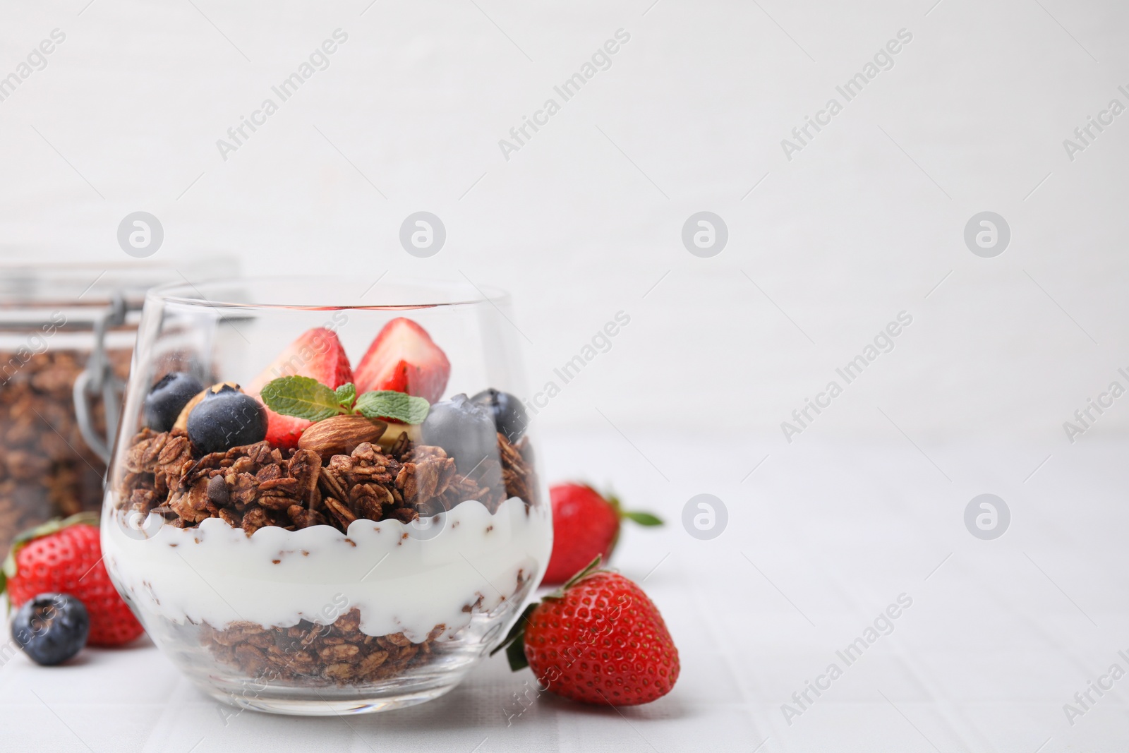 Photo of Tasty granola with berries, yogurt and mint in glass on white tiled table, space for text