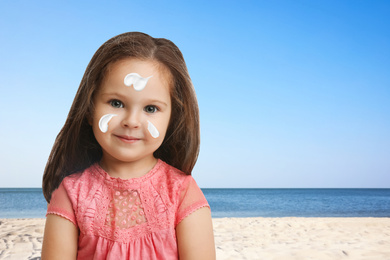 Adorable little girl with sun protection cream on face at sandy beach, space for text 