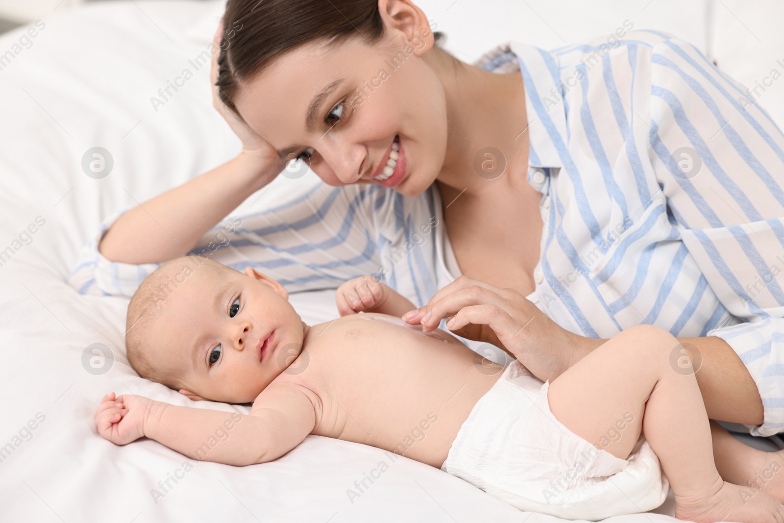 Photo of Happy young woman applying body cream onto baby`s skin on bed