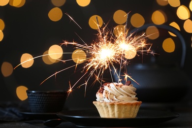 Photo of Cupcake with burning sparkler on black table against blurred festive lights