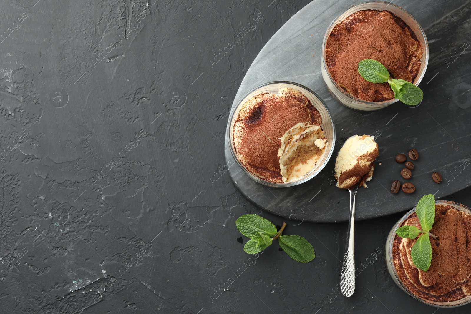 Photo of Delicious tiramisu in glasses, spoon, mint leaves and coffee beans on black table, top view. Space for text