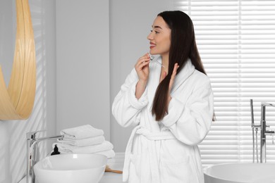 Photo of Happy young woman applying essential oil onto hair near mirror in bathroom