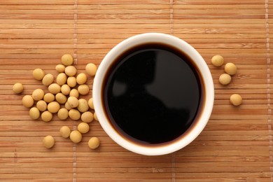Soy sauce in bowl and soybeans on bamboo mat, flat lay