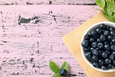 Photo of Bowl of tasty fresh bilberries and green leaves on old pink wooden table, flat lay. Space for text