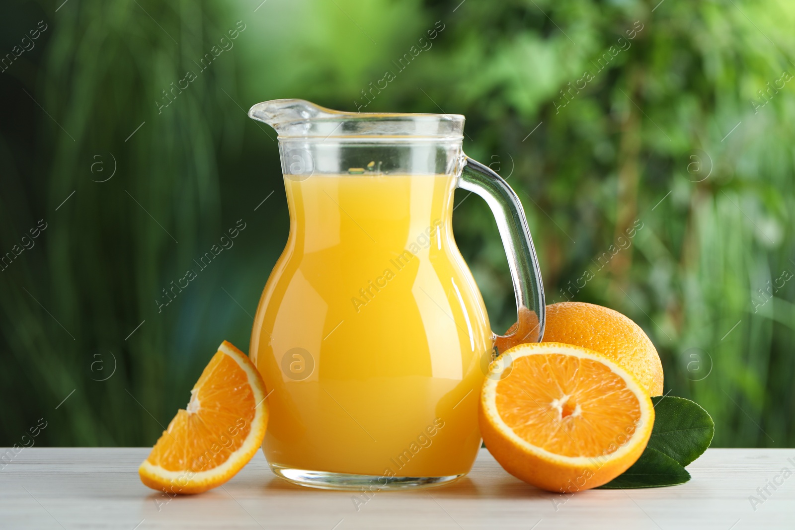 Photo of Tasty orange juice in glass jug, fresh fruits and green leaves on white wooden table outdoors