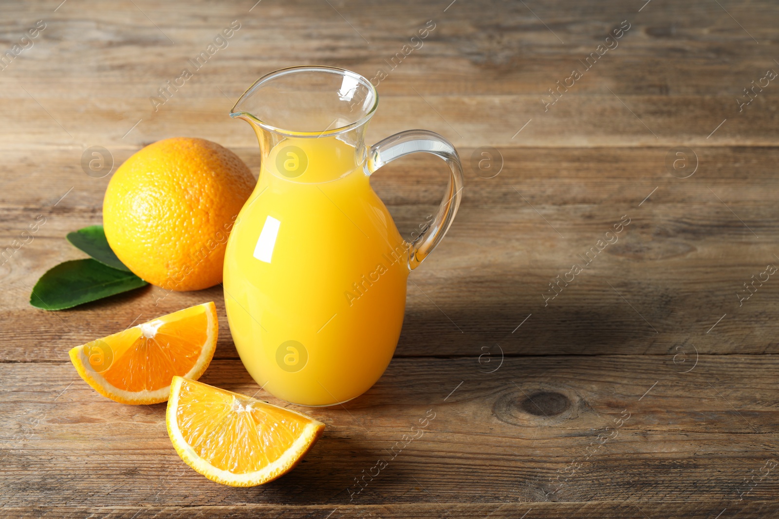 Photo of Tasty orange juice in glass jug, green leaves and fresh fruits on wooden table, space for text