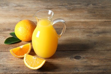 Photo of Tasty orange juice in glass jug, green leaves and fresh fruits on wooden table, space for text