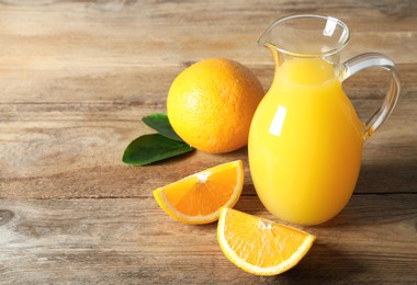 Tasty orange juice in glass jug, green leaves and fresh fruits on wooden table. Space for text