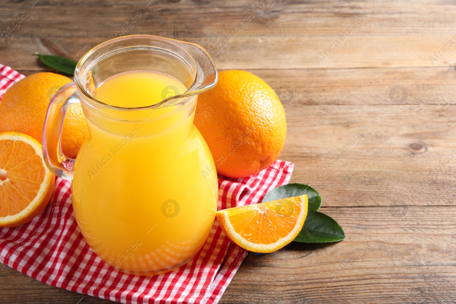 Photo of Tasty orange juice in glass jug, fresh fruits and green leaves on wooden table, space for text