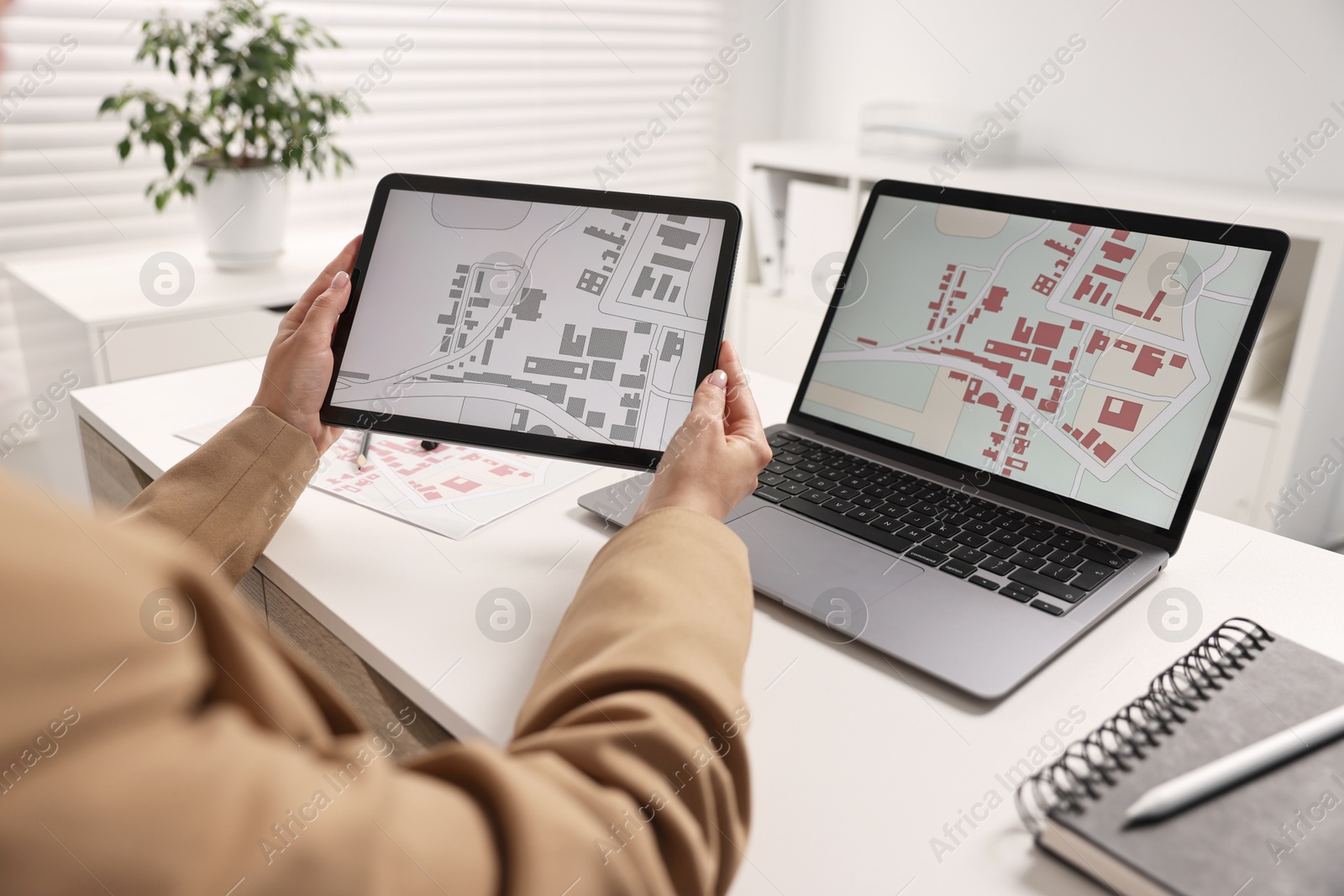 Photo of Cartographer working with cadastral maps on devices at white table in office, closeup