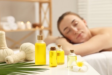 Photo of Aromatherapy. Woman relaxing on massage couch in spa salon, focus on bottles of essential oils, herbal bags and plumeria flower