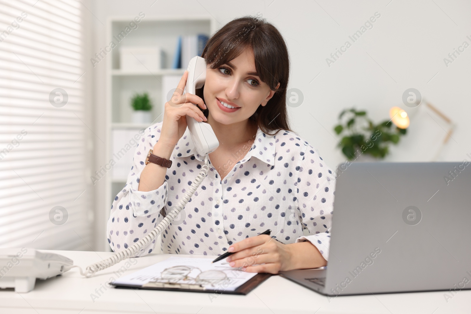 Photo of Smiling secretary talking on telephone at table in office