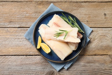 Photo of Pieces of raw cod fish, rosemary and lemon on wooden table, top view