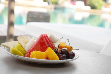 Plate with fresh fruits on table near sun lounger, space for text. Luxury resort with outdoor swimming pool