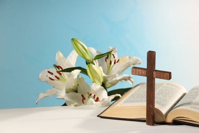 Wooden cross, Bible and beautiful lily flowers on white table against light blue background, space for text. Religion of Christianity