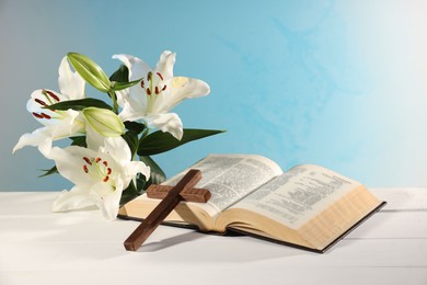 Photo of Wooden cross, Bible and beautiful lily flowers on white table against light blue background. Religion of Christianity
