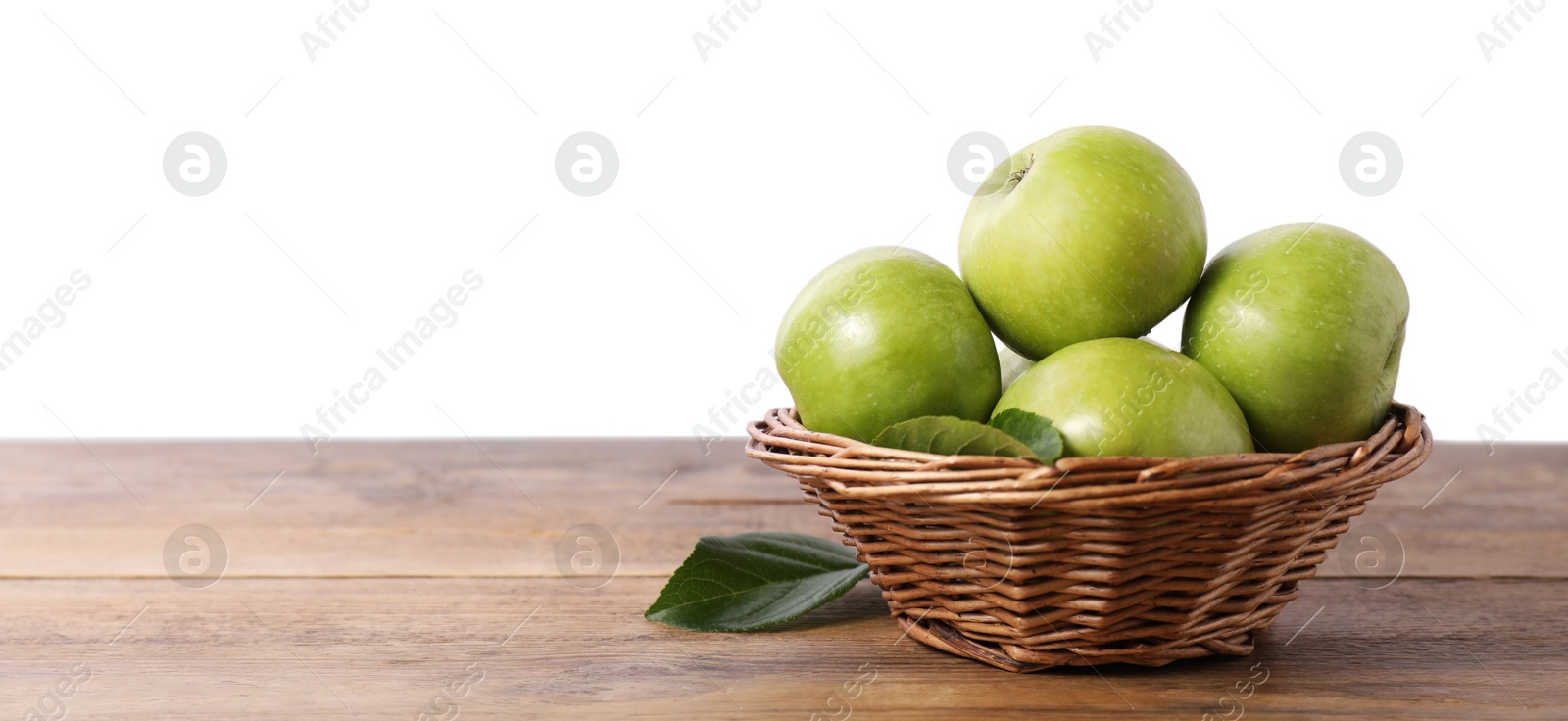 Photo of Ripe green apples in wicker basket on wooden table against white background. Space for text