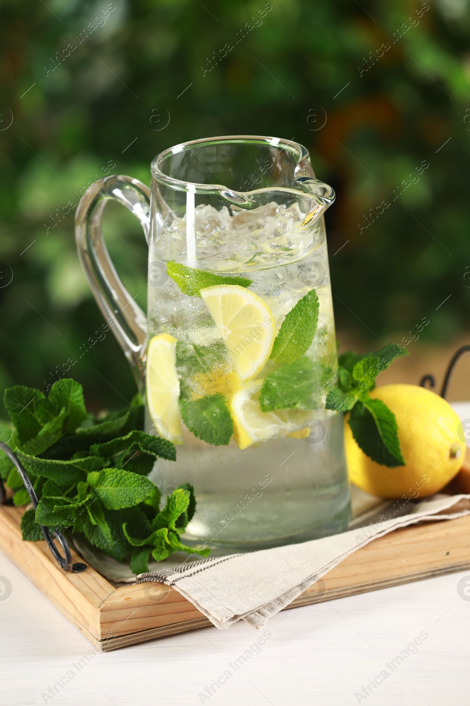 Photo of Refreshing lemonade with mint in jug and citrus fruit on light table against blurred green background