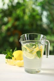 Refreshing lemonade with mint in jug and fruits on light table against blurred green background