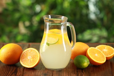 Photo of Refreshing lemonade with citruses in jug among fruits on wooden table against blurred green background